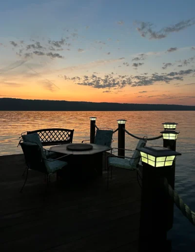 Patio furniture on a dock at sunset with lit lanterns and a calm lake in the background.