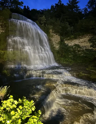 Illuminated waterfall cascading over rocky ledges surrounded by green foliage at twilight.